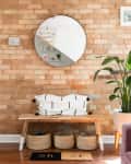 Entryway with brick wall, round mirror, wooden bench, woven baskets, and a large potted plant.