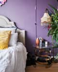 Bedroom with purple wall, upholstered headboard, yellow pillow, glass side table, vintage lamp, and potted plant.