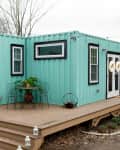 Turquoise shipping container home with a wooden deck, white doors, black trim, and outdoor seating with a small table.