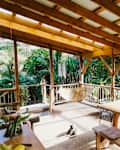 Covered wooden patio with a hammock, dining table, and a cat, surrounded by lush greenery.