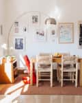 Dining room with wooden table, woven chairs, red high chair, wall art, and potted plants by a window.