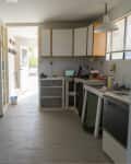 Outdated kitchen with white cabinets, fridge, stove, and cluttered countertops under a window with blinds.