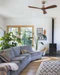Bright living room with gray sofa, wicker chairs, wood stove, large plants, and framed art, featuring natural light from windows.