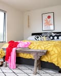 Bedroom with a yellow bedspread, patterned pillows, wooden bench, and colorful clothes. Large window and abstract wall art.