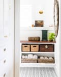 Hallway with woven baskets, rolled towels, decorative boxes, a plant, and a wall sconce on patterned tile floor.