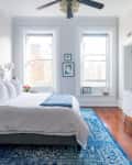 Bright bedroom with a gray tufted bed, blue rug, white dresser, and wall art, featuring large windows and a ceiling fan.
