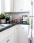 Bright kitchen corner with white cabinets, stainless steel sink, dish rack, potted plants, and colorful bowls on the counter.