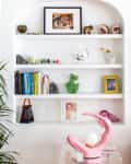 White wall niche with shelves displaying books, framed photos, sculptures, and decor items. Pink lamp on glass table.
