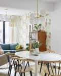Dining room with floral wallpaper, round table, wooden chairs, green sofa, and hanging plants.
