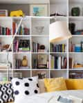Bookshelf with colorful books, modern lamp, yellow sofa, and patterned cushions in a cozy living room.