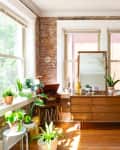 Mid-century modern dresser with plants, mirror, and decor in a sunlit room with brick walls and wooden floors.