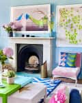 Colorful living room with blue walls, vibrant artwork, green coffee table, pink flowers, and a white fireplace.