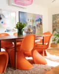 Dining room with orange chairs, wooden table, pink pendant light, and colorful abstract wall art.