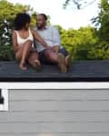 Couple sitting on a small house roof, surrounded by trees, enjoying a sunny day.
