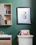 A bathroom painted dark green with a small vanity, a mirror opening to reveal a medicine cabinet, and a framed drawing above the white toilet.