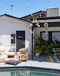 Poolside patio with white sofa, wicker chairs, potted plants, and string lights against a white building.