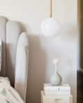 Gray upholstered headboard, geometric patterned bedding, white pendant light, and small vase on stacked books.