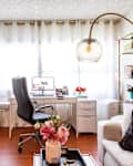 Home office with white desk, black chair, large plant, teepee, and gold floor lamp. Bookshelf and sofa with black pillow.