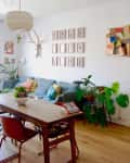 Living room with a wooden dining table, colorful chairs, gray sofa, wall art, plants, and a large window.