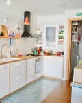 Bright kitchen with white cabinets, wooden countertops, open shelves, colorful dishes, and a patterned blue floor mat.