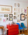 Gallery wall with diverse framed art, a wooden table with a potted plant, red drawers, and two blue chairs.