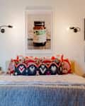 Bedroom with colorful patterned pillows, wall sconces, framed art, and a small side table with books.