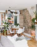 Open-concept living room with exposed brick wall, fireplace, plants, dining table, and kitchen with bar stools.