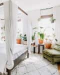 Canopy bed with white curtains, surrounded by potted plants, green velvet chair, and a dresser in a bright bedroom.