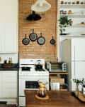 Cozy kitchen with white cabinets, brick wall, hanging pans, and a wooden table with a coffee maker and plants.