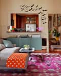Cozy living room with gray sofa, orange polka dot blanket, pink rug, and bat wall decals near a kitchen entrance.