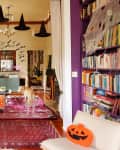Purple living room with Halloween decor, hanging witch hats, pumpkins, bookshelves, and a dog on a red rug.