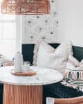 Cozy breakfast nook with a round marble table, wicker pendant light, patterned pillows, and a potted plant by the window.