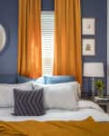 Bedroom with blue walls, orange curtains, round mirror, and a bed with white and blue pillows, flanked by two lamps.