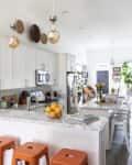 Bright kitchen with white cabinets, marble countertops, orange stools, hanging lights, and plants near a dining area.