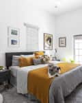 Cozy bedroom with a gray bed, mustard throw, cat, wall art, plants, and a side table with a lamp.