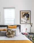 Bedroom with a dark headboard, yellow and white pillows, eclectic wall art, and vintage side tables with decor items.