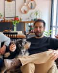 Two men sitting on a brown leather sofa with two dogs in a cozy living room with plants and wall decor.