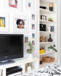 Bedroom with a TV, framed photos on the wall, and a white shelf with books and decor items. Dark door in the background.