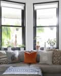 Gray sofa with assorted pillows, surrounded by potted plants, under large windows in a bright living room.