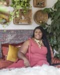 Woman in a pink dress sitting on a bed with colorful pillows, surrounded by plants and woven wall decor.