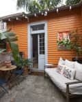 Orange patio with a white door, potted plants, blue chairs, a small table, and a bench with colorful pillows.