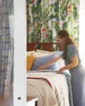 Woman making a bed with tropical leaf curtains, yellow and blue pillows, and a wooden dresser with a wine bottle and flowers.