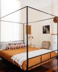 Canopy bed with orange bedding, patterned pillows, and a decorative fireplace in a vintage-style bedroom.