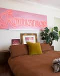 Bedroom with brown bedding, a yellow pillow, a pink "Romance" sign, a plant, and a white dresser with framed photos.