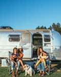 Family sitting outside a silver Airstream trailer with a dog, surrounded by trees and a parked truck.