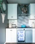 White kitchen with blue stove, patterned backsplash, teal pot, and floral wreath above.