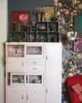 White cabinet with glassware and plates, surrounded by plants, art, and floral wallpaper, next to a red armchair.