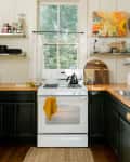 Cozy kitchen with white stove, black kettle, yellow towel, and colorful wall art. Shelves hold dishes and jars.