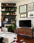 Cozy living room with a beige sectional sofa, black shelves with plants, framed art, and a TV on a wooden dresser.