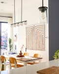 Modern dining area with wooden table, tan chairs, geometric wall art, pendant lights, and large plants by a window.